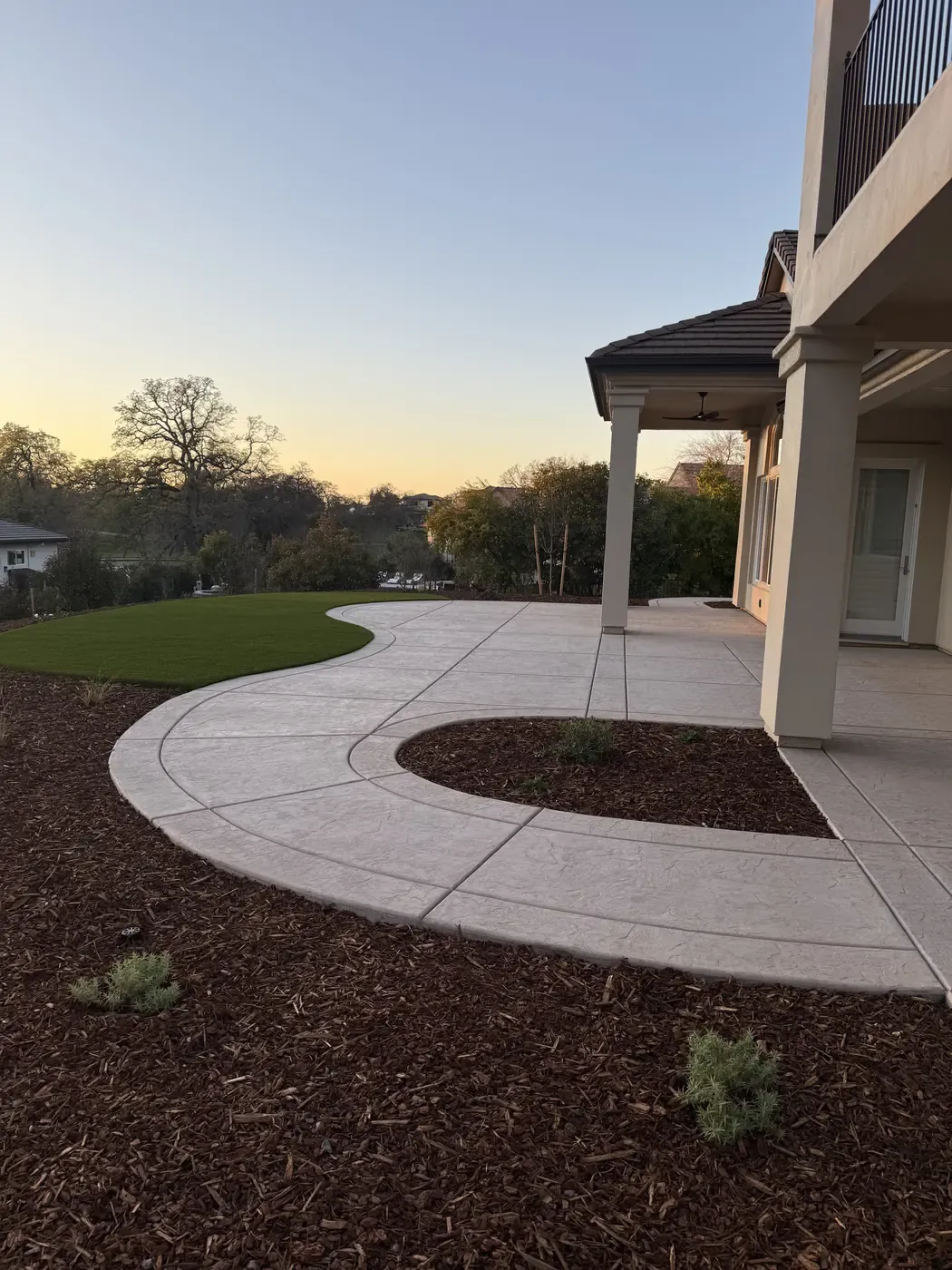 Curved stamped concrete patio and walkway wrapping a planting bed behind a two story home at sunset