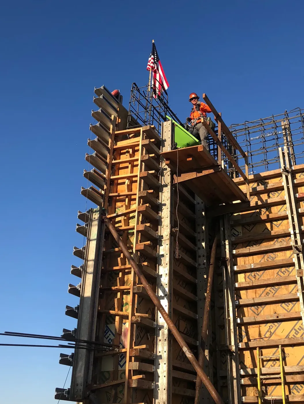 Colton in a hard hat and safety gear atop tall concrete column forms on a commercial job, with an American flag on the rebar
