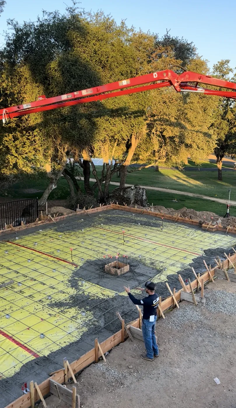 CJR crew member directing a concrete pump boom over a reinforced slab ready for a pour, framed by oak trees