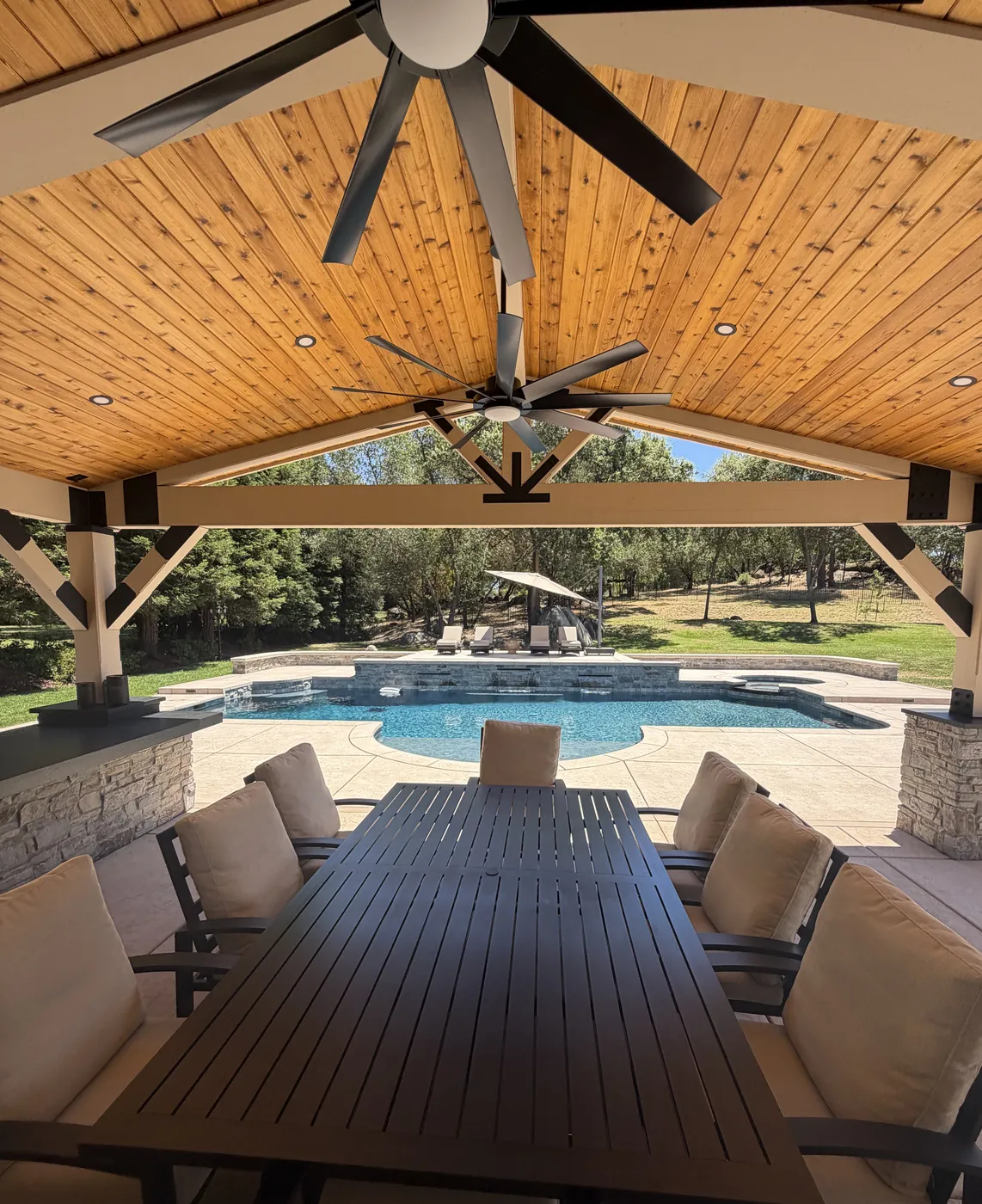 View from a cedar ceiling pavilion over a dining table toward a pool with sheer descents and lounge chairs