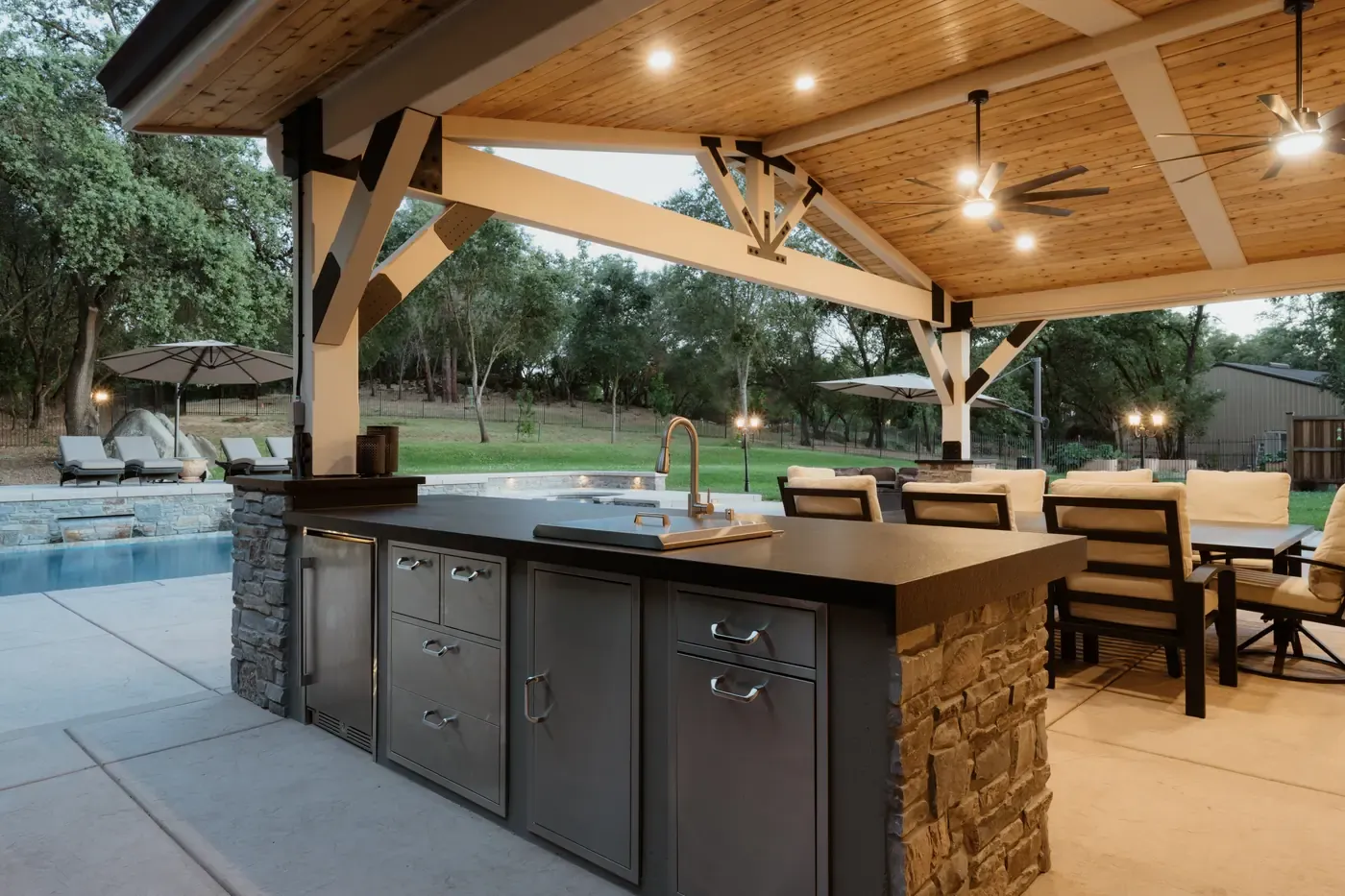 Covered outdoor kitchen with a stone island, ceiling fans and poolside seating at dusk