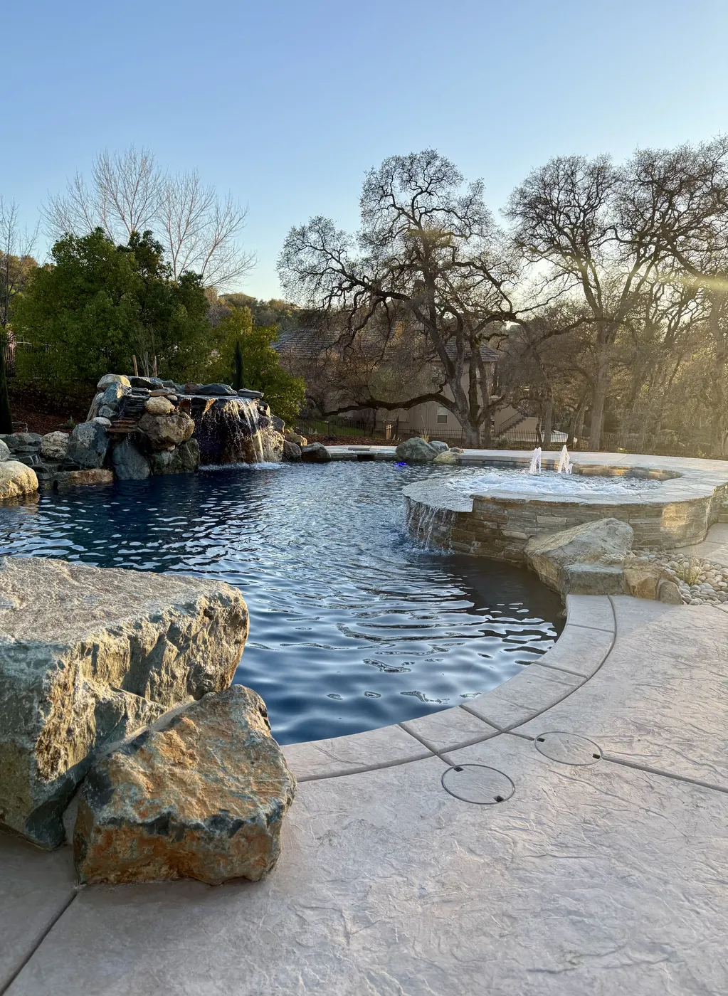 Natural boulder pool with a rock waterfall, raised spa with bubblers, and stamped concrete decking under mature oaks