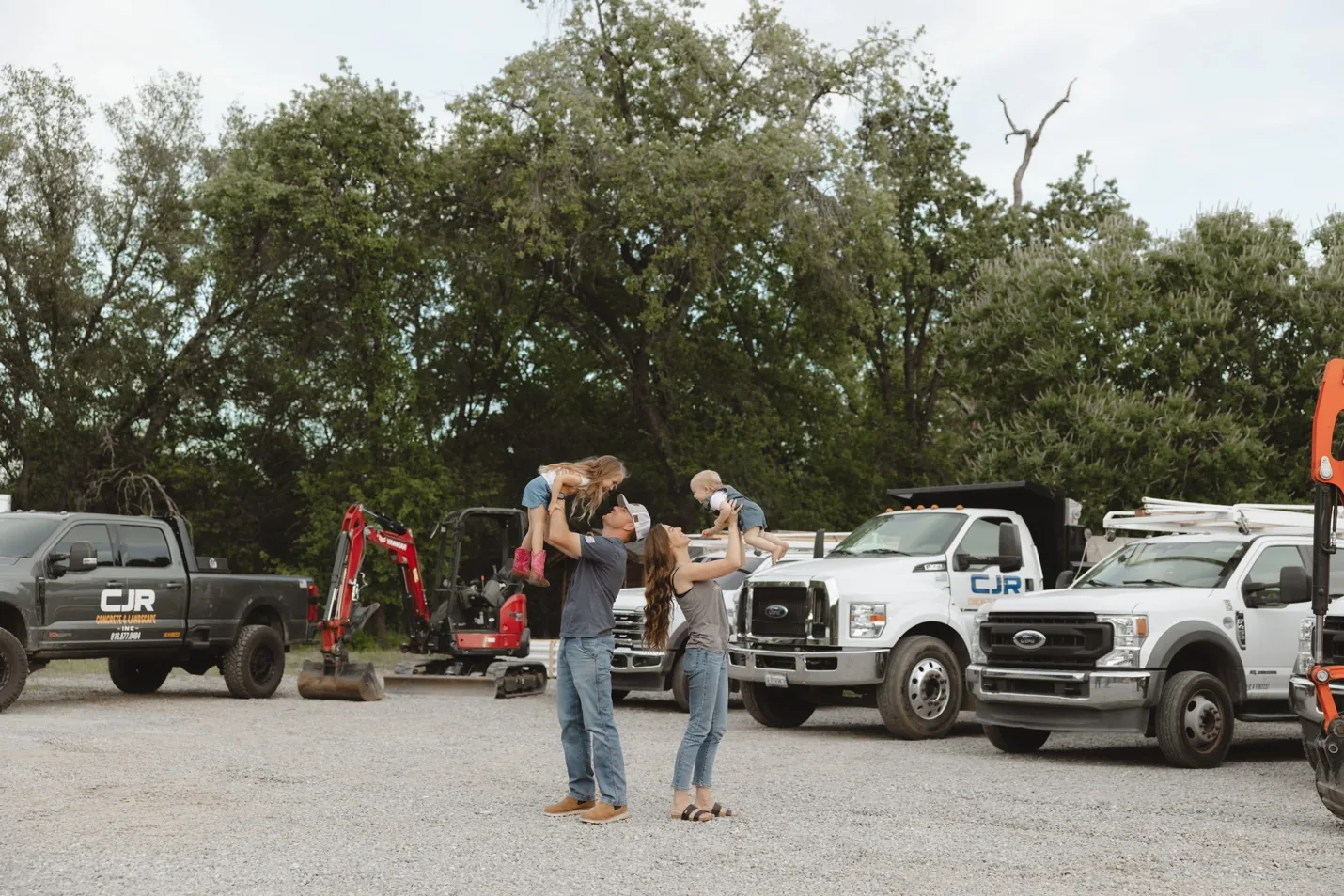 Colton and Paige Ray lifting their two children in the air in front of the CJR trucks and excavator in Loomis